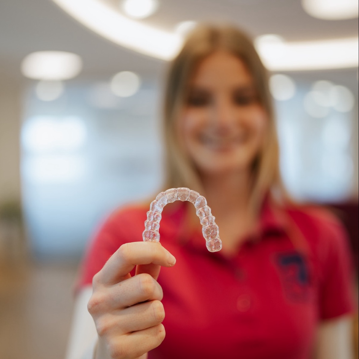 Eine durchsichtige Aligner-Schiene in der Hand, dahinter lächelt eine Mitarbeiterin der Praxis